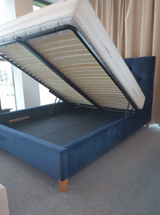 tapicerowane łóżko w stylu glamour z wysokim, pikowanym zagłówkiem i miękką pianką. Posiada duży pojemnik na pościel z łatwym dostępem (podnośniki gazowe). Stabilne, na wysokich dębowych nogach. Dostępne w wielu tkaninach