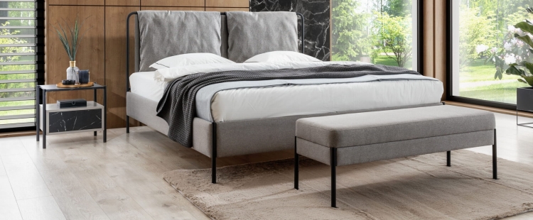 Łóżko tapicerowane z lekką ramą z okrągłych metalowych rurek, miękkie poduszki na zagłówku i stylowe czarne nóżki. Komfort i nowoczesność w jednym meblu.